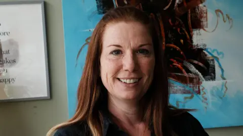 A head and shoulders image of a woman wearing a black top. She is looking into the camera. She is standing in front of some artwork which is on the wall.