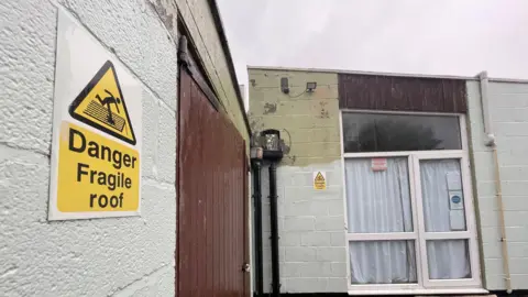 A yellow and white sign says "danger fragile roof", it is on an outside wall. IN the background is a window with curtains drawn.