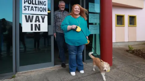 PA Woman with ginger hair in turquoise top and jeans with a light coloured small dog at a polling station 