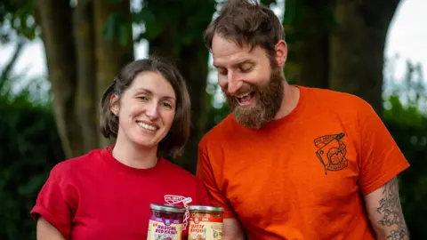 The Crafty Pickle Co Madi Myers and Arthur Serini pose holding jars of their sauerkraut and kimchi products. Madi wears a red top with The Crafty Pickle Co logo and has a brown bob hairstly. Arthur wears an orange tshirt with The Crafty Pickle Co logo and has short brown hair and a full brown beard. 