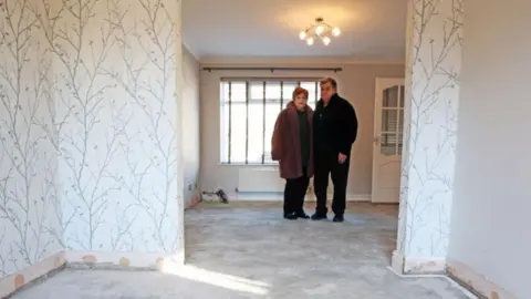 A man and a woman are stood together in their house which has a flood-damaged floor