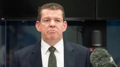 PA Media Rhun ap Iorwerth speaking into a microphone. He wears a dark suit, green patterned tie and white shirt.