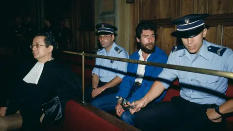 Getty Images Georges Abdallah se sienta esposado en un banco judicial entre dos policías. Lleva jeans azules, una chaqueta azul, una camisa blanca, y tiene una barba gruesa y marrón y bigote