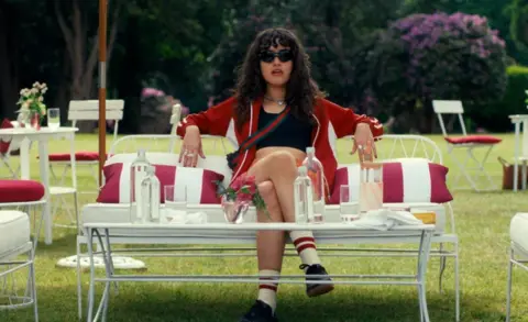 Amazon Asha Banks as Noah sits on a sofa in a garden wearing sunglasses and a red hoodie