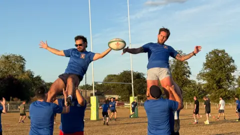 Elliot Deady/BBC Two rugby players are lifted into the air by their teammates. Tehy are all wearing matching blue kits. Several players are playing a game behind them.