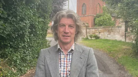Scott Spencer Scott wears a check shirt and jacket and is standing outside a church.