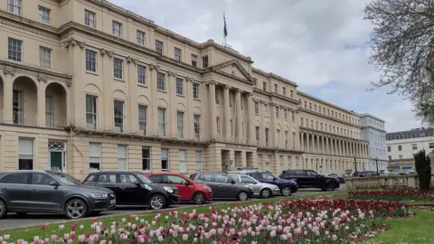 A large Regency style building with cars parked outside and a manicured green lawn with pink and purple tulips growing on it. 