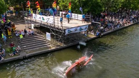 PA Media A large scaffolding structure with rainbow-coloured flags is overlooking the harbourside in Bristol. In the water is a homemade flying contraption which looks like a small red plane with wings. Crowds of people are watching from the side.
