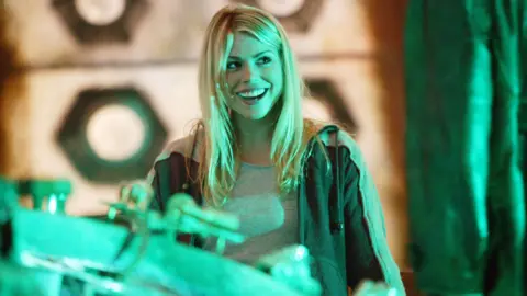 BBC Billie Piper on the set of Doctor Who. She is looking to the right of the photo and is smiling. Her blonde hair is shoulder-length and she has a grey t-shirt and dark grey jacket on. 