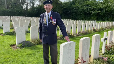 Olivia Richwald/BBC An old man wears a jacket covered in medals and rests his hand on a white headstone