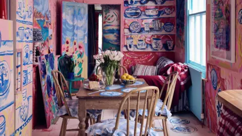 Alun Callender A dinning room with pink walls, features hand painted pictures of Dutch blue and white pottery associated with Holland. A wooden table and chairs is in the centre of the picture with flowers and cake on it.
