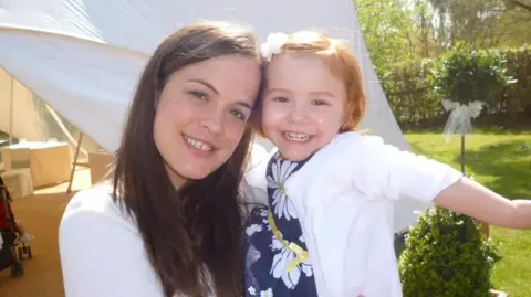 Grace Kelly Childhood Cancer Trust a woman in a white top is smiling and holding a young young who is wearing a blue dress with white flowers and a white cardigan