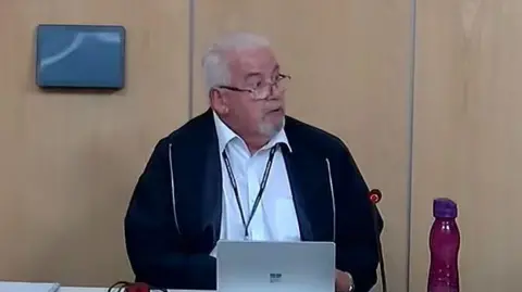 A man sitting at a table in a council meeting room. He is looking to his left. He has white hair and a white goatee and is wearing glasses. He is wearing an unzipped blue jacket over a white shirt. His laptop is open in front of him and there is a purple water bottle to his left.