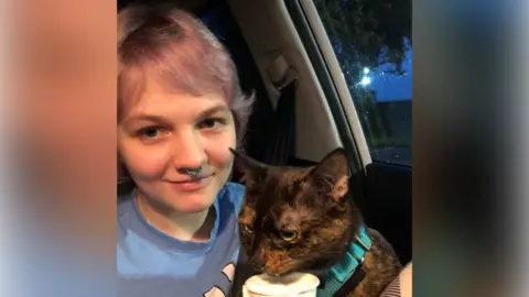 Family handout A head and shoulders shot of Libby Mycroft with a black and brown cat on her lap. The cat has a blue collar and is eating from a cup. Libby is wearing a blue T-shirt and has short, light purple hair and a nose ring. She is sitting in a passenger seat of a car and is smiling at the camera.