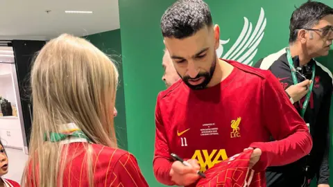 Shauna O’Carolan Girl with blonde hair wearing a red and black Liverpool Football Club jacket. She has blue eyes. She is getting a Liverpool football shirt signed by Liverpool player Mo Salah who is wearing the team's red colours