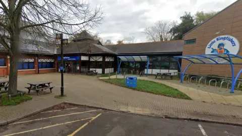 Bricked pool building to the left with a reception area in the centre of the photo. A large circular sign saying Little Kingfishers is on the right with an empty bike rack in front of it. There is also a litter bin and two picnic benches in the foreground.