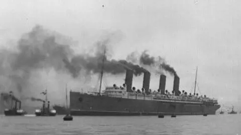Tyne & Wear Archives A back and white image of a ship on the water, with dark smoke rising out of its four funnels. The base of the ship is darker that the floors on top. There are smaller vessels on the water beside it, with tall masts rising from them.