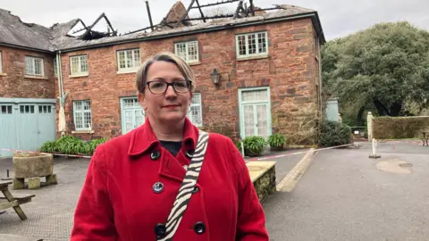 Kate Ewert wearing a red coat standing in front of the fire-damaged building. . She has blonde hair and black glasses. The roof damage is shown with the trusses visible.