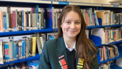 Picture of student Evelyn smiling at the camera. Evelyn has dark brown hair and brown eyes. She's wearing her school uniform of dark green blazer, blue jumper and white blouse. She is standing in a library setting with rows of books behind her on a book case.
