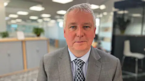 A man with a grey suit and light blue shirt looks at the camera in front of an office.