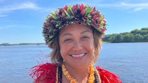 A smiling Dr Melissa Taitano, in traditional brightly coloured head dress and necklaces, stands on the shores of the River Foyle