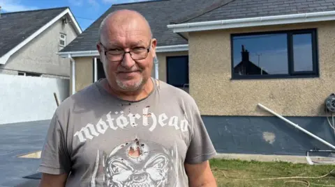BBC William standing in front of what looks to be a beige pebbledash one-storey home with a driveway in front. He has glasses and a short grey beard and short hair, and is wearing a grey Motorhead t-shirt.