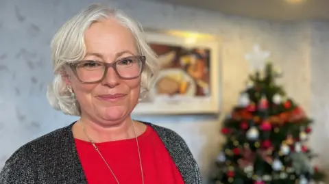 Nickia is wearing a bright red top and a dark gray cardigan stands indoors near a decorated Christmas tree adorned with red, gold, and white ornaments. The background features a light-colored patterned wall and a framed picture.