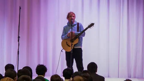 Yayasan Ed Sheeran Ed Sheering di atas panggung di depan siswa sekolah. dia memegang gitar dan mengenakan hoodie biru muda dan celana panjang gelap