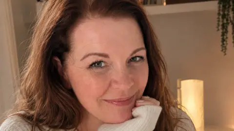Samantha Thomas Samantha Thomas is pictured with long auburn hair wearing a cream knitted jumper, resting her chin on one hand and looking towards the camera. Warm indoor lighting, framed photographs and houseplants are visible on shelves in the softly blurred background.