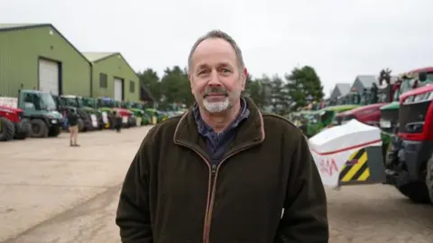 Shaun Whitmore/BBC A man with grey short hair and a hair goatee beard smiles at the camera. He is wearing a dark green coat and a blue jumper underneath. Tractors are packed behind him either side in a row.