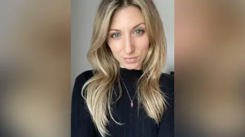 A woman with long hair and a dark coloured top. She is looking into the camera in the selfie.