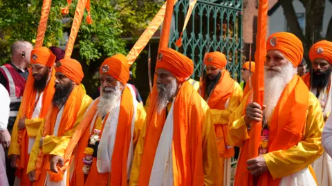 Men in orange turbans and scarves dressed in white robes lead a procession through the streets. The are carrying poles wrapped in orange fabric. The men have long beards.