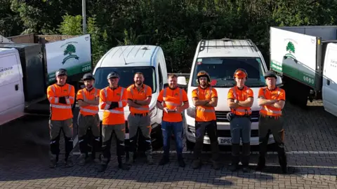 DLM Tree Services DLM Tree Services team stood in in front of white vans wearing orange hi vis uniforms.