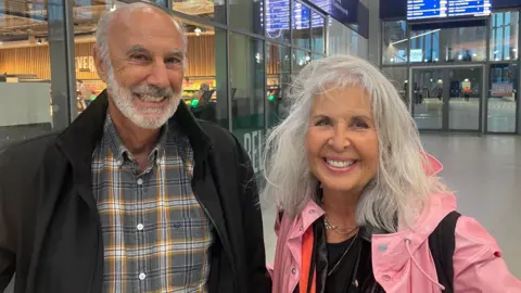 Michael and Iitta looking at the camera, Michael is wearing a black jacket with a grey and orange check shirt. he has white hair and beard. Iitta wearing a pink jacket, she has silver hair.