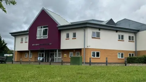 Andrew Turner/BBC Mile Cross Primary School, featuring yellow brickwork on the lower half, white rendering to the upper storey, and a grey corrugated metal roof. There is a gable, painted purple, which bears the name in silver letters, Mile Cross Primary School. In the foreground is a lawn, some black bollards and a green utility box.