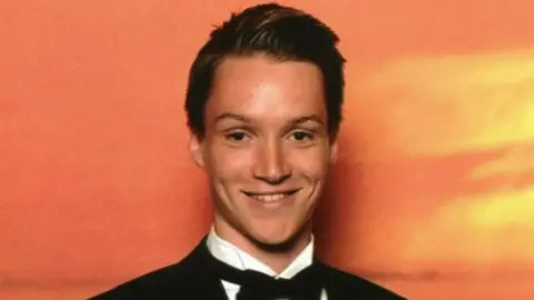 Lisa Wilson Tom Wilson smiles at the camera, he is wearing a tuxedo and is stood in front of an orange background.