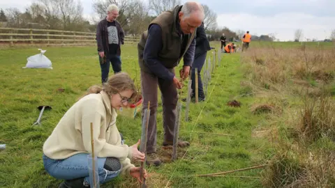 RSPB A woman and man fixing wire into the ground. The woman is crouched down. She has light brown hair in a pony tail. She is wearing a beige jumper and blue jeans. The man is stood up. He is wearing brown trousers, a long sleeve dark blue jumper and a khaki green body warmer.