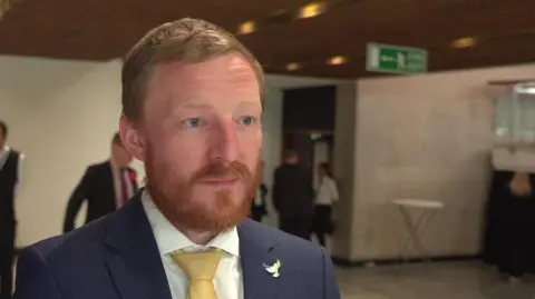 Colin Ferguson. He has red hair and a beard. He is wearing a blue suit with a yellow tie.