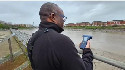 A man in a dark fleece jacket with short cropped grey and black hair stands with his back to the camera looking down a hand held air quality tester. He is holding the tester above a metal railing on the promenade above a brown river with wide muddy banks and a housing development with a row of four storey red brick buildings above the furtherst bank. 