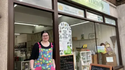 A woman with black hair and glasses wears a black top and a colourful apron with leaves on it as she stands in the doorway of her refill shop. Large bottles of liquid with big pumps can be seen inside the shop and there are scarecrows, shelving, and signs in the window.