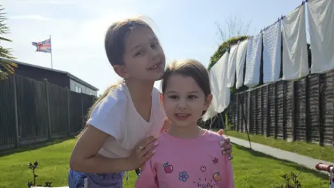 Supplied Two girls, one in a white t-shirt and one in a pink t-shirt in a sunny back garden
