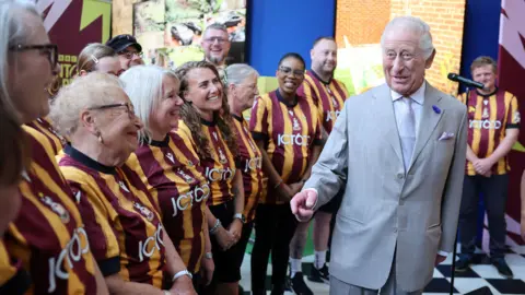PA Media King Charles wearing a light grey suit stands in front of a choir of people wearing claret and amber-striped Bradford City football shirts.