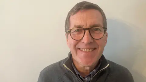 Mark Hunter, the leader of Stockport Council, smiles wearing a grey zipped jacket and shirt, with tortoise shell glasses, while stood against a beige-painted wall. 