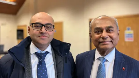 Shariqua Ahmed/BBC Nabil, a man, with a bald head, wearing a white shirt and a blue tie and a grey coat, standing next to Mohssin, a man, with short grey hair, wearing a blue striped suit and a blue tie, smiling for camera.
