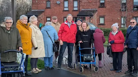 Brian Wernham About a dozen elderly men and women stand with Councillor Brian Wernham outside a bus stop and a row of houses in a square. They are wearing colourful coats and two people have mobility aid walkers. 