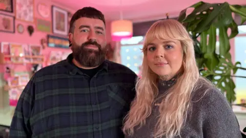 A man with dark hair and a blue and green tartan shirt stood next to a woman with blonde hair wearing a grey jumper.
