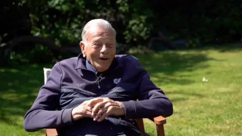 Dickie Bird sitting down in a wooden chair in his garden, smiling in the sun.