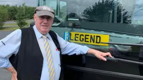 A man in a cap, waistcoat, shirt and tie standing in front of a coach. There is a number plate in the window with "legend" printed on it.