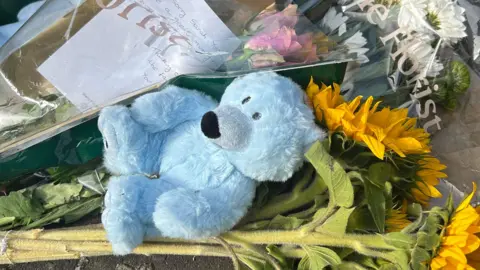 BBC A close up of blue teddy bear, a bunch of sunflowers and floral bouquets with handwritten notes. They are among items placed in tribute to Donaghadee murder victim Sarah Montgomery.