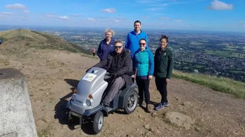 A man sits on a grey buggy on top of the Malvern Hills while a group of four others stand beside him wearing coats and walking clothing.